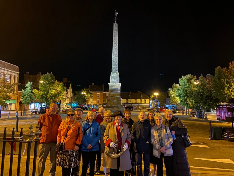 Members of the group stand around the Ripon Hornblower in the market square at night,
with the Ripon obelisk monument standing tall behind them