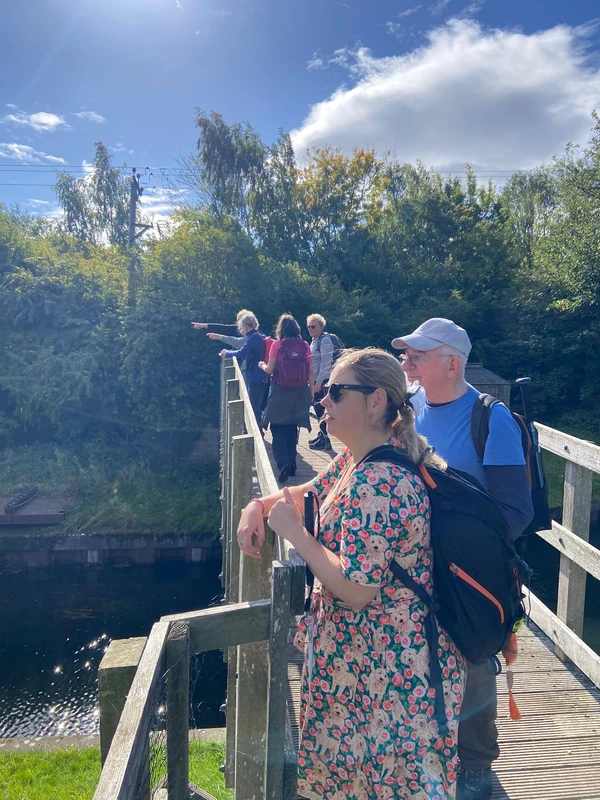 The group stands on a wooden canal bridge basked in sunlight,
some pointing to things in the distance out of shot