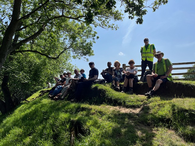 The group stop at the top of a hill in front of a fence, partly shaded from an overhanging tree, with all sitting except for one member in a high-vis jacket