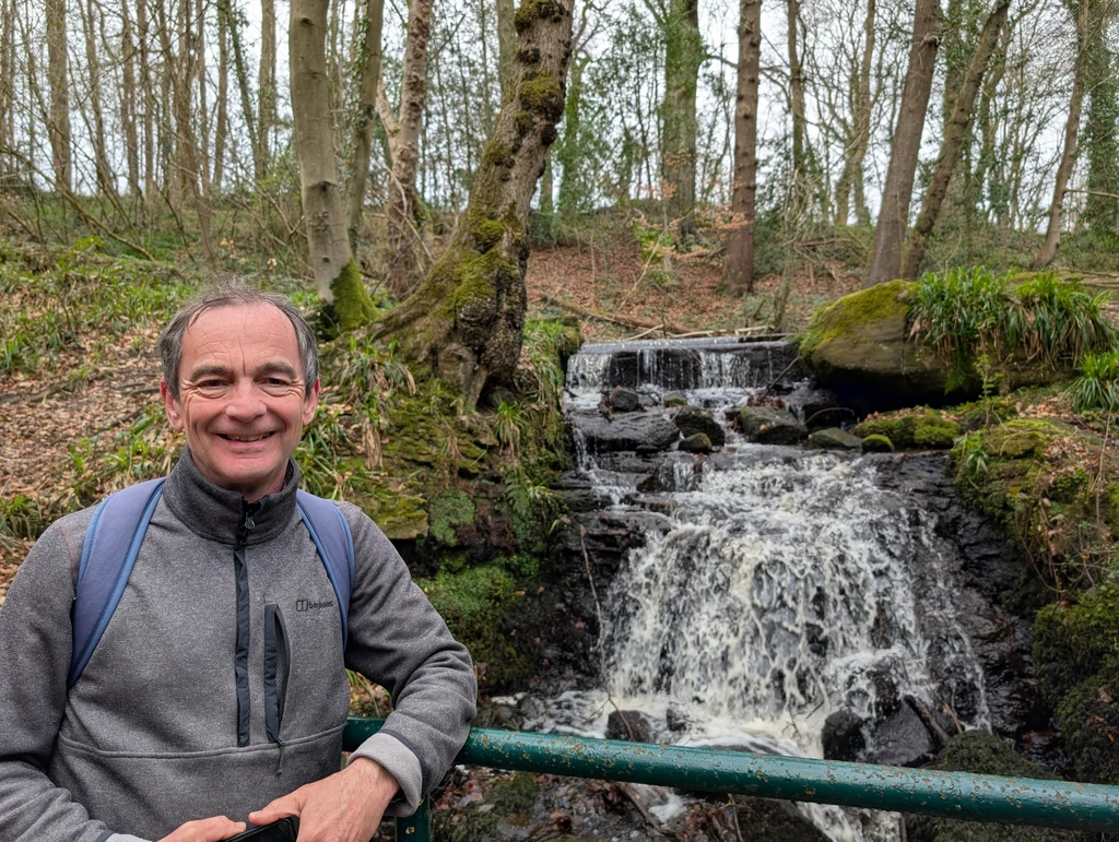 A member of the group leans against the railing of a woodland bridge above a stream
