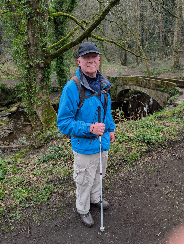 A member of the group stands on a dirt footpath in a wooded area near a bridge